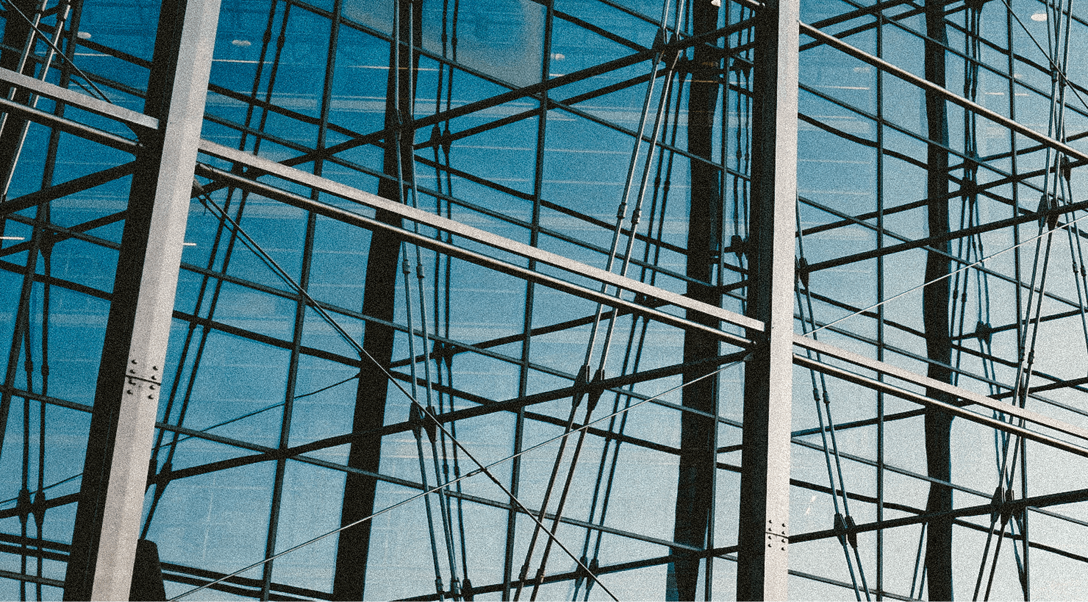 Close-up view of a modern glass building facade with intersecting metal beams and cables, reflecting a blue sky and creating a complex geometric pattern.